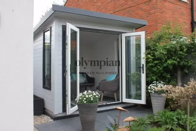 An 8ft x 8ft painted and insulated garden building with single-sloping roof and UPVC doors, between house wall and fence, in a landscaped setting