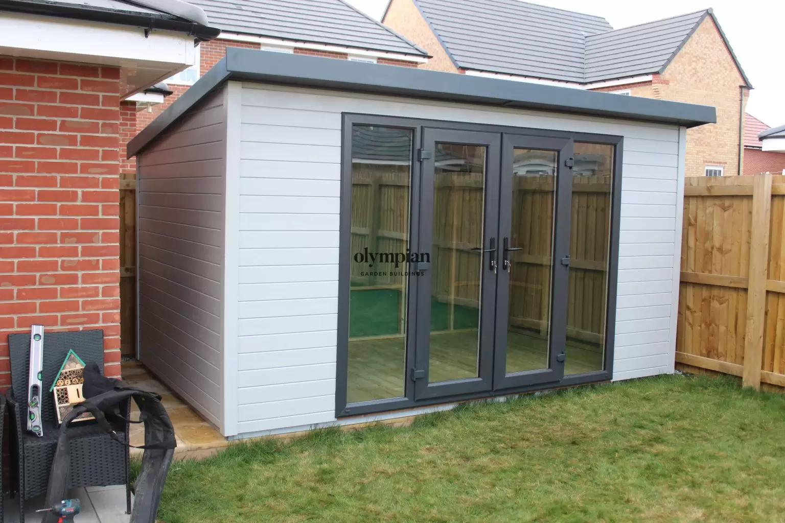 A 13ft 6inch x 8ft painted garden building with single-sloping roof and UPVC doors in the corner of a garden