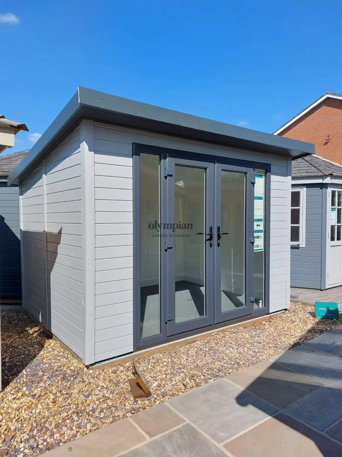 A 10ft x 8ft painted garden building with single-sloping roof and UPVC doors, surrounded with other garden buildings between landscaped Indian stone paving and stones