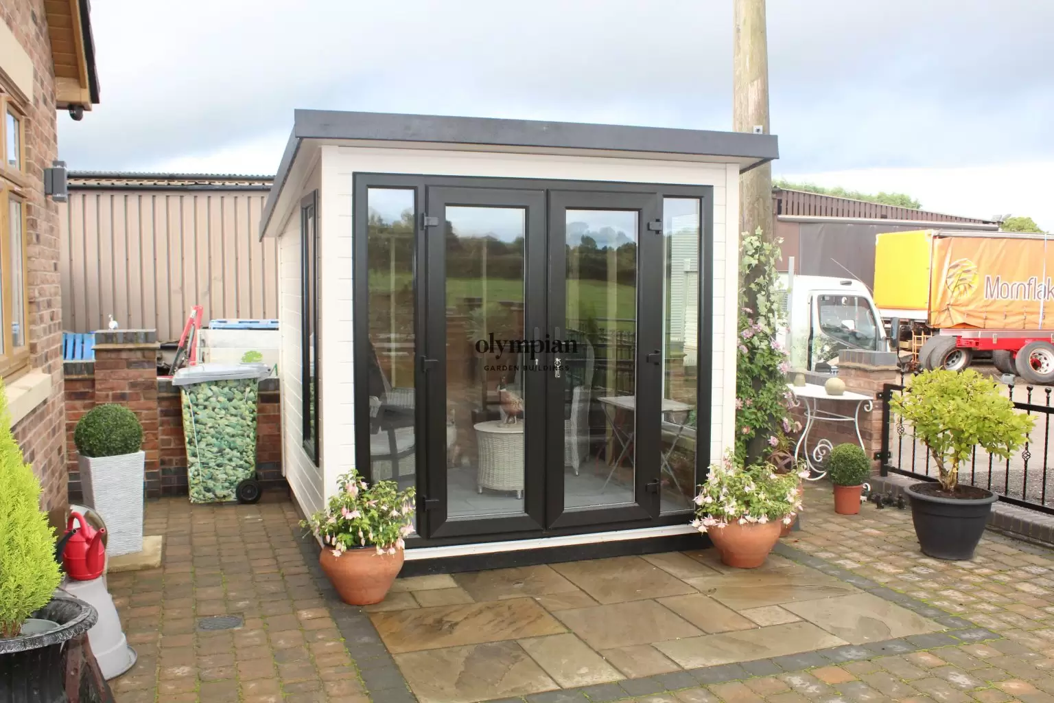 An 8ft x 8ft painted garden building with single-sloping roof and UPVC doors, in an open paved garden