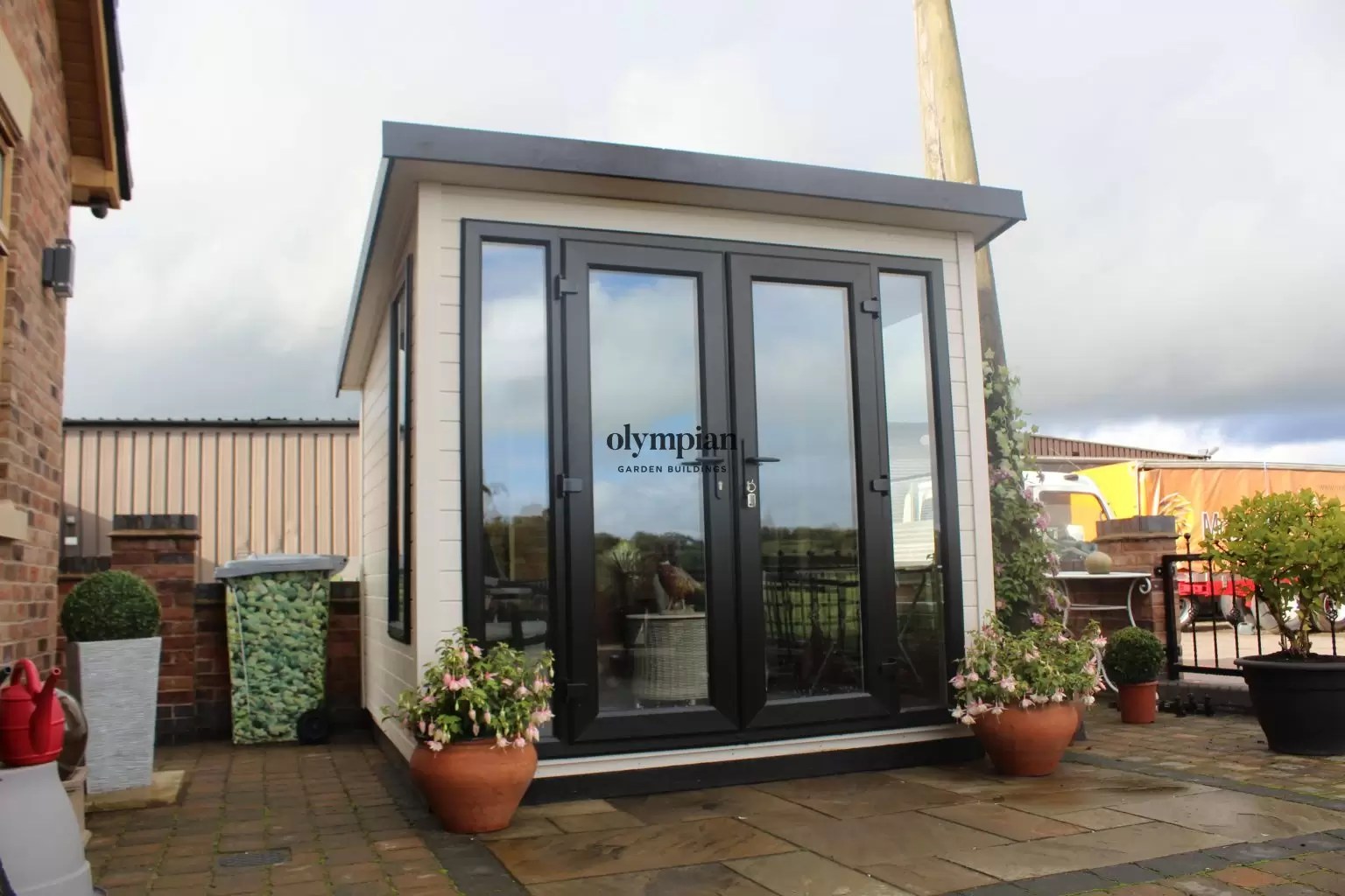 An 8ft x 8ft painted garden building with single-sloping roof and UPVC doors, in an open paved garden